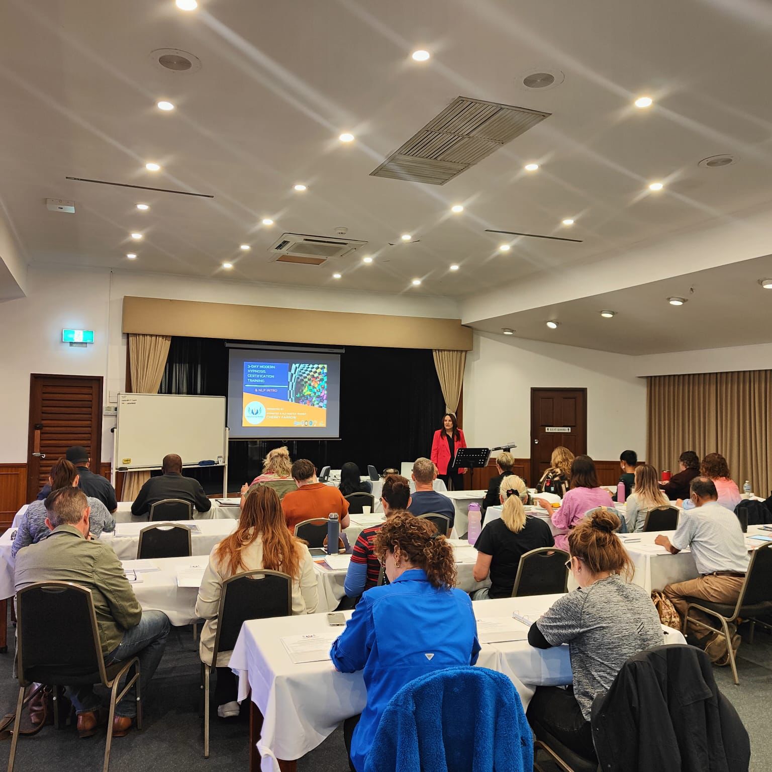 Conference or classroom setting with attendees at tables and a presenter at the front